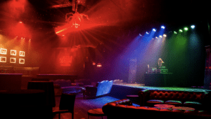Empty nightclub interior with colorful lighting. A DJ stands at a booth on the right. Tables, chairs, and a couch are visible under red, blue, and green lights.
